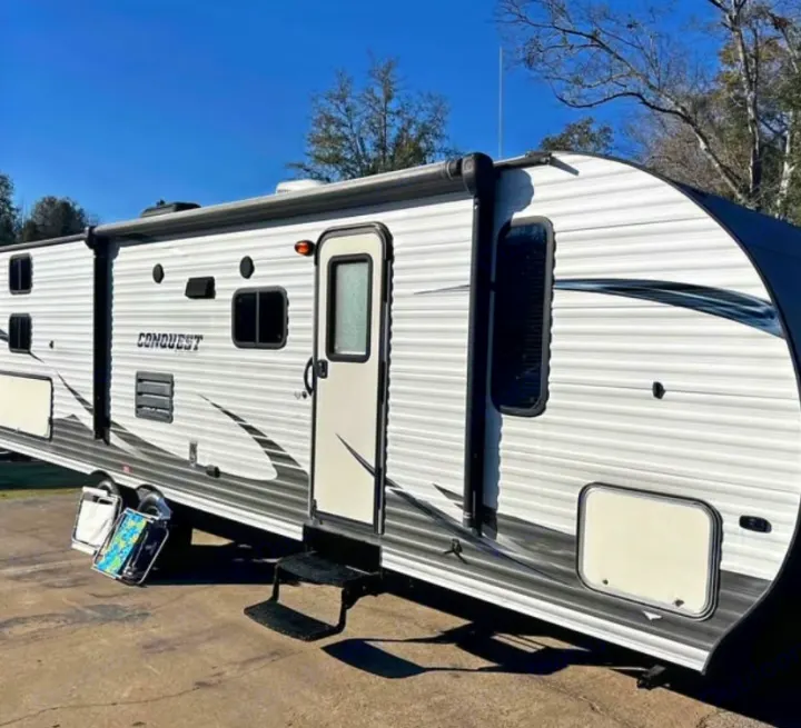 A white and gray travel trailer, a Conquest RV, with an awning extended, is parked outdoors. Perfect for your next RV rental.. Gulf Stream Conquest 2017