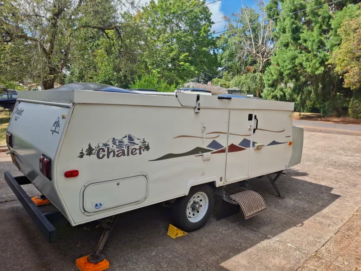 White Chalet pop-up camper, perfect for an RV rental. Parked on concrete with stabilizing jacks and mountain decals.. Chalet Chalet Popup Trailer 2006