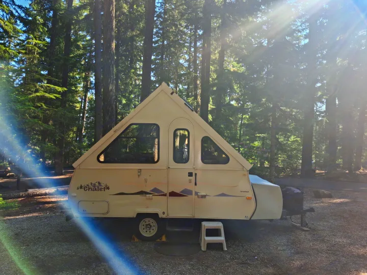 A beige A-frame pop-up RV rental is parked on gravel at a sunny forest campsite, surrounded by tall trees.. Chalet Chalet Popup Trailer 2006
