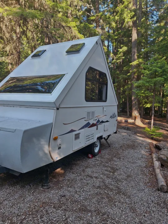 White A-frame RV rental parked on a gravel campsite amidst tall forest trees.. Chalet Chalet Popup Trailer 2006