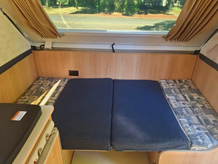 An rv rental's cozy interior, featuring a bed/seating with blue and patterned cushions, a counter, and window.. Chalet Chalet Popup Trailer 2006