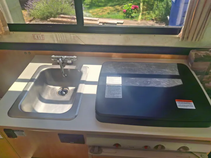 Compact kitchen in an rv rental, featuring a stainless steel sink, covered stovetop, and window with an "EXIT" sign.. Chalet Chalet Popup Trailer 2006