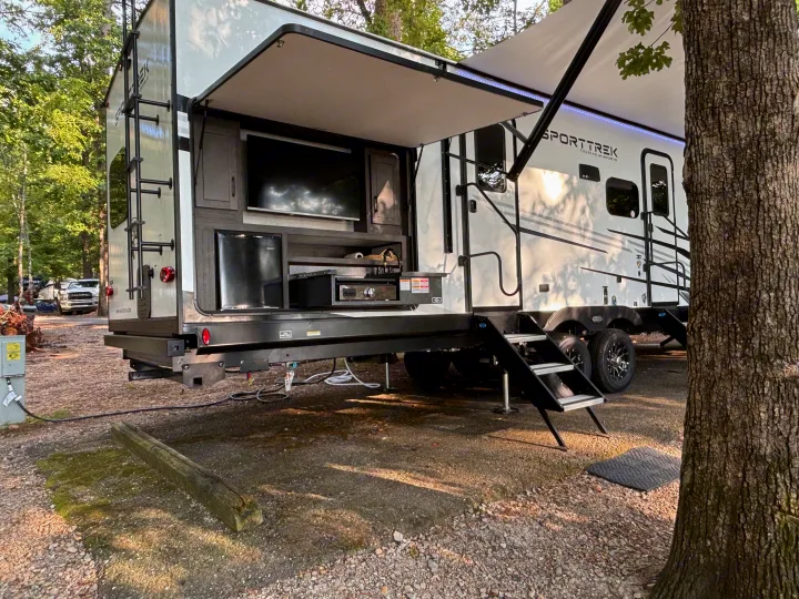 An RV rental, a SportTrek model, parked at a campsite with its outdoor kitchen (TV, fridge, sink) open and awning extended.. Venture Rv SPORTTREK TOURING EDITION 2025
