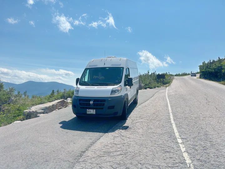A white RV rental camper van is parked on a scenic mountain road under a bright blue sky, with distant peaks visible.. Dodge B Van 2018