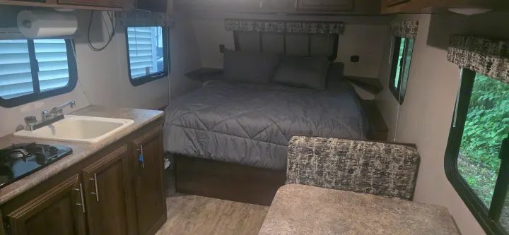 RV rental interior featuring a bed, kitchen area with sink/stove, and dinette. Windows show nature and nearby structures.. KZ Sportsman Classic 180 QB 2018