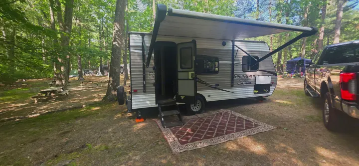 RV rental: Travel trailer set up at a wooded campsite with an awning, outdoor rug, picnic tables, and pickup truck.. KZ Sportsman Classic 180 QB 2018