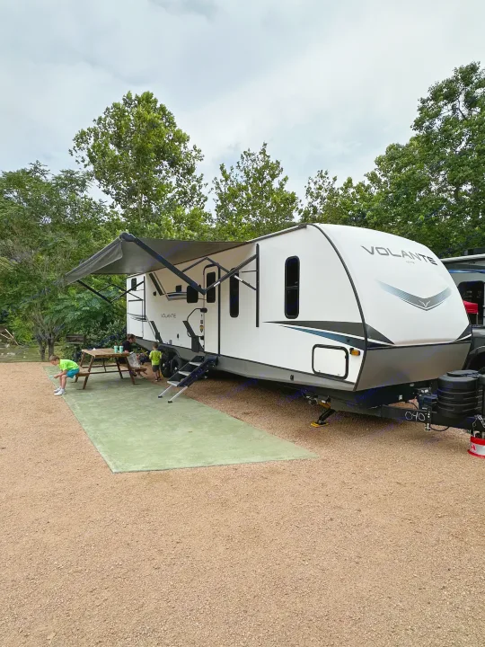 Family enjoys an RV rental at a scenic campsite with an extended awning & picnic table.. Crossroads RV Volante 2023