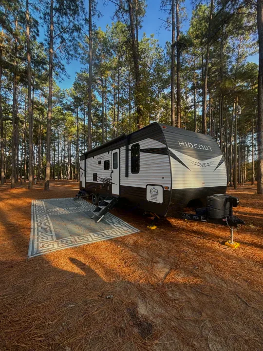 An RV rental travel trailer is set up with an outdoor rug at a sunny pine forest campsite.. Keystone Hideout 2020