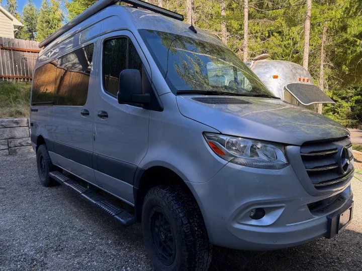 A silver Mercedes-Benz Sprinter camper van, ready for RV rental, parked on gravel with trees & an Airstream trailer.. Thor Tranquility 2022