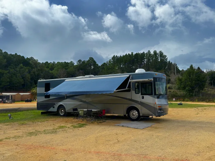 Large gray RV rental with extended awning and outdoor seating at a gravel campground under cloudy skies.. Itasca Horizon 2002