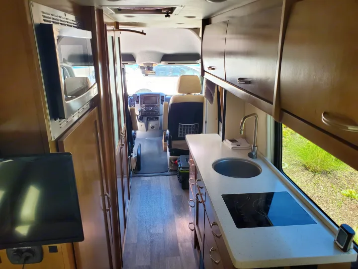 Inside this modern RV rental: a compact kitchen with sink/cooktop, microwave, TV, and the driver's cabin view.. Coachmen Galleria 2019