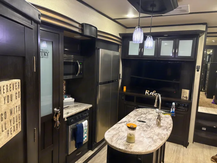 View of a modern RV rental interior: dark wood kitchen cabinetry, stainless appliances, island with sink, and living area with TV.. Grand Design Momentum Toy Hauler 2015