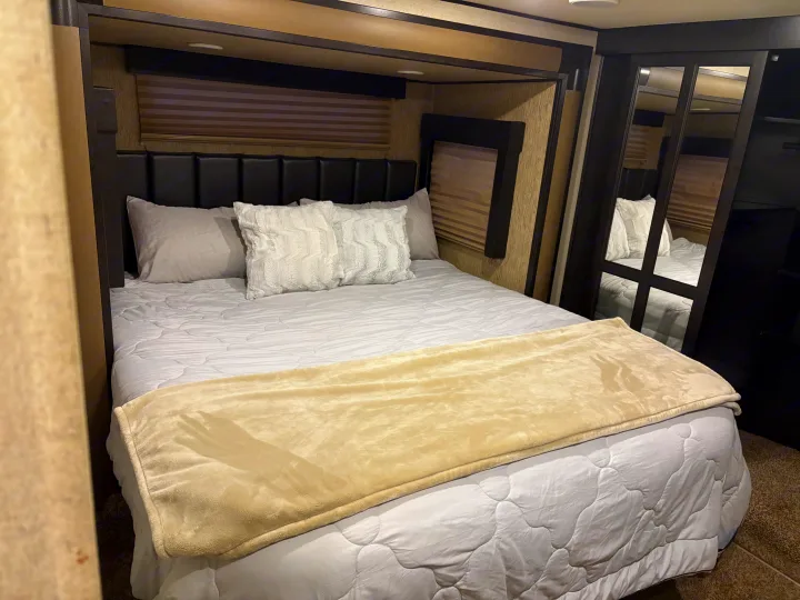 Comfortable RV rental bedroom with a plush bed, modern headboard, window blinds, and mirrored closet doors.. Grand Design Momentum Toy Hauler 2015