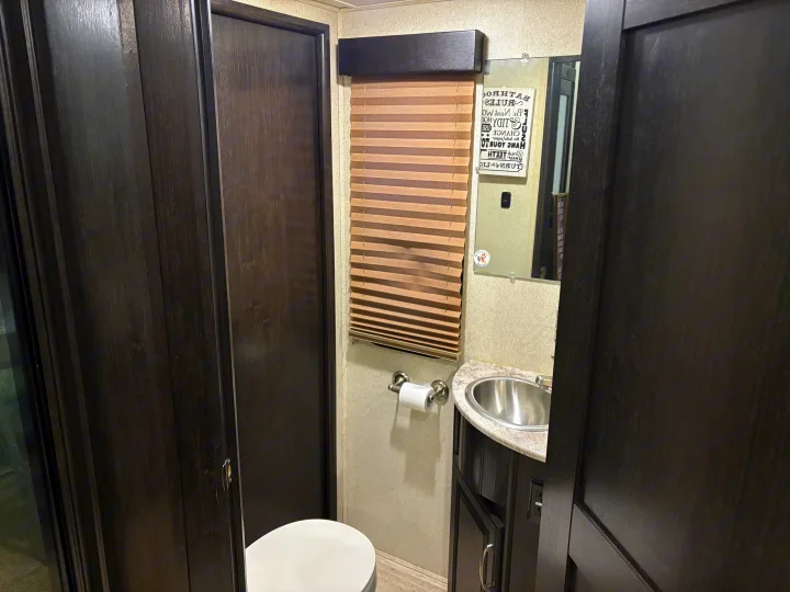 Inside an RV rental bathroom, featuring dark wood doors, a toilet, sink, and mirror with a 'Bathroom Rules' sign.. Grand Design Momentum Toy Hauler 2015