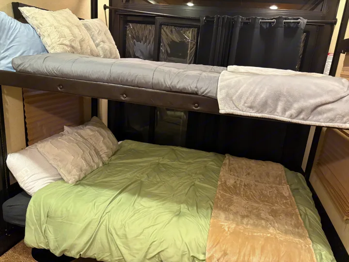 RV rental bunk beds feature grey and green bedding. Windows behind them have black curtains and light-colored blinds.. Grand Design Momentum Toy Hauler 2015
