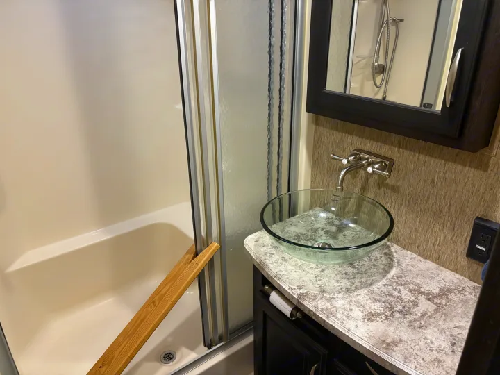 Compact RV rental bathroom: white shower/tub with glass door, vessel sink, and a wooden plank propped inside.. Grand Design Momentum Toy Hauler 2015