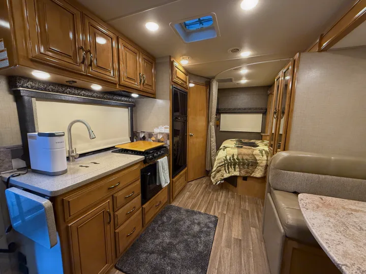 Bright rv rental interior featuring a full kitchen with a Keurig, stove, and sink. Includes a comfortable bed and dinette space.. Thor Quantum 2018
