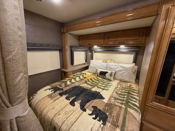 Cozy, bear-themed bedroom interior of an RV rental, featuring wooden cabinetry, headboard, and windows with blinds.. Thor Quantum 2018
