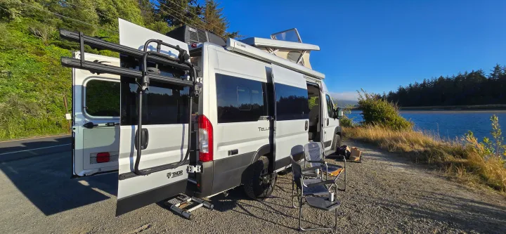 This rv rental, a white camper van with pop-top, chairs, and bike rack, is parked by a scenic river for camping.. Thor Tellaro 2024