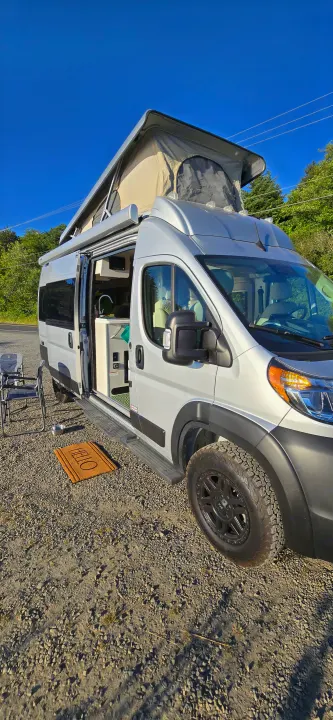 Explore in this rv rental: a silver pop-top camper van with open door, interior view, and outdoor chairs on gravel.. Thor Tellaro 2024
