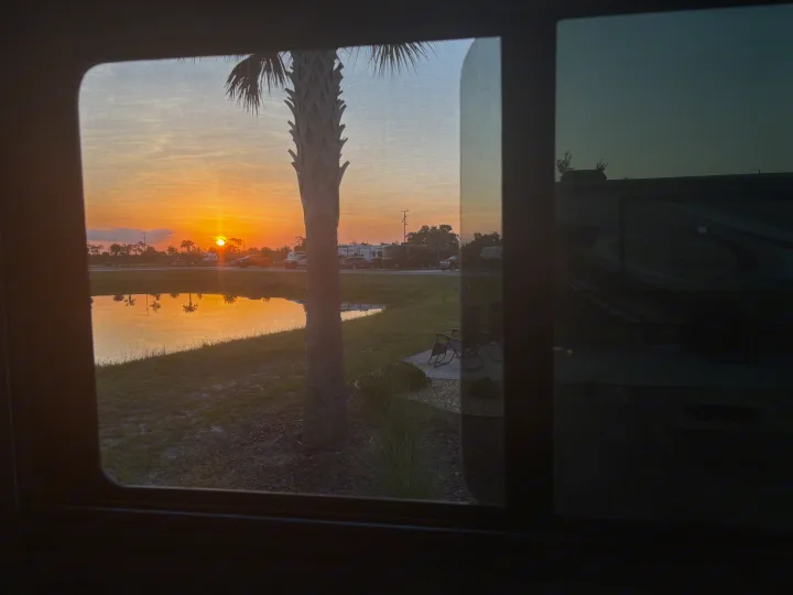 Vibrant sunset through an RV window. A palm tree stands by a pond reflecting orange light, with other RVs nearby. Ideal for an RV rental.. 1FDWE3FNXSDD14715 Cross trail 22xgf 2025