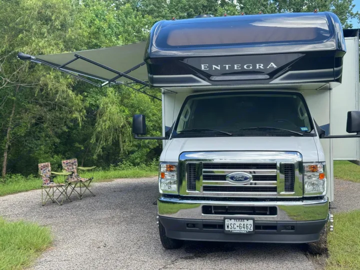 White Entegra Ford RV rental parked on a gravel path with awning extended. Two camping chairs nearby for outdoor enjoyment.. Entegra Coach Odyssey 2021