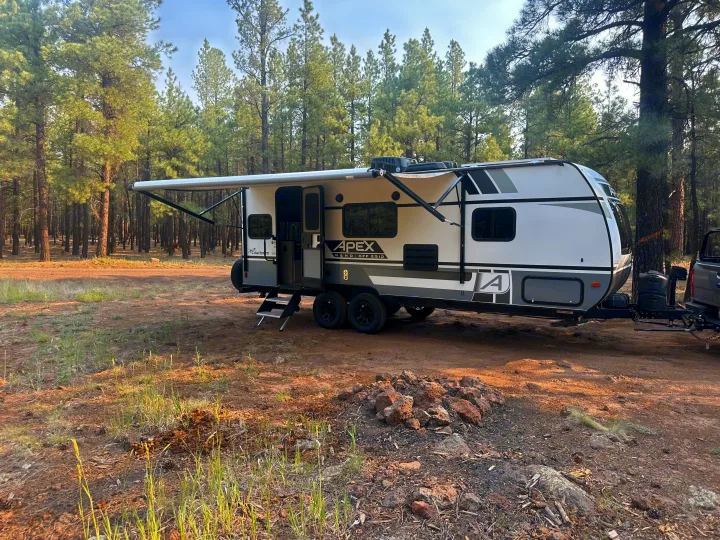 A modern white Apex Nano off-grid RV rental travel trailer is parked in a pine forest campsite.. Coachmen Apex Nano - Off Grid 2022