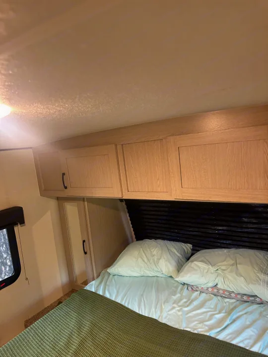 RV rental bedroom featuring a cozy bed with light blue sheets, overhead light wood cabinets, and a window.. Coachmen Apex Nano - Off Grid 2022