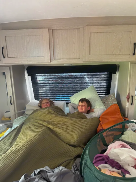 Smiling boys relax comfortably in their RV rental bunk, covered by a green blanket. Perfect for family travel and cozy adventures.. Coachmen Apex Nano - Off Grid 2022