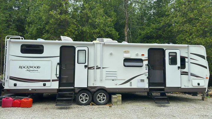 White Rockwood Signature RV rental parked on gravel with trees, showing its two entry doors and full side profile.. Forest River Rockwood Signature Ultra Lite 2015
