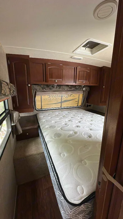 RV rental bedroom with a white quilted mattress, dark wood cabinets, and large window offering natural light, ready for travel.. Forest River Rockwood Signature Ultra Lite 2015