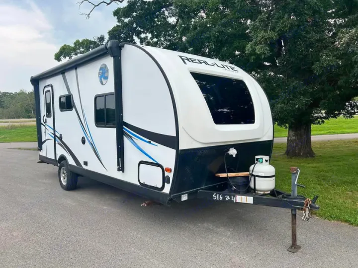 White & black Real-Lite travel trailer, ideal for an RV rental, sits on asphalt. Features a large front window & propane tank.. Palomino Real Lite 2019