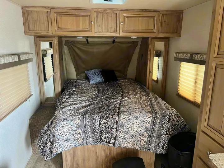 Cozy RV rental bedroom featuring a bed with patterned comforter, overhead cabinets, and compact wardrobe space.. Palomino Real Lite 2019