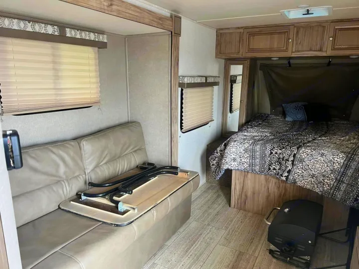 RV rental interior features a comfortable bed with patterned bedding, a convertible sofa, and overhead cabinets. A portable grill sits on the floor.. Palomino Real Lite 2019