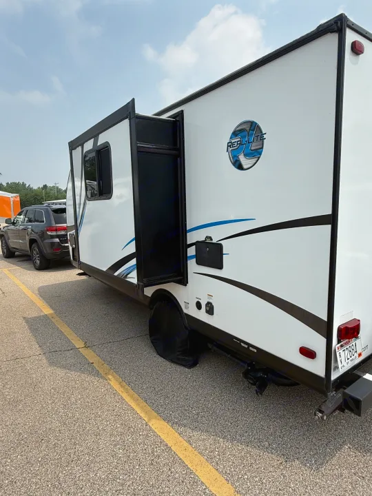 This white REAL-LITE RV rental is parked with its side door open, hitched to a dark SUV on a sunny day. Wisconsin plate visible.. Palomino Real Lite 2019