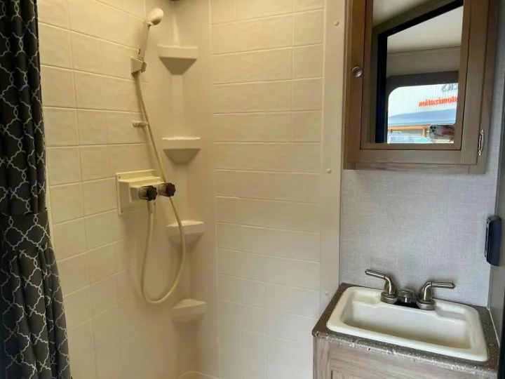 Inside an RV rental: a bright, compact bathroom with a white-tiled shower, handheld sprayer, small sink, and mirrored cabinet.. Palomino Real Lite 2019