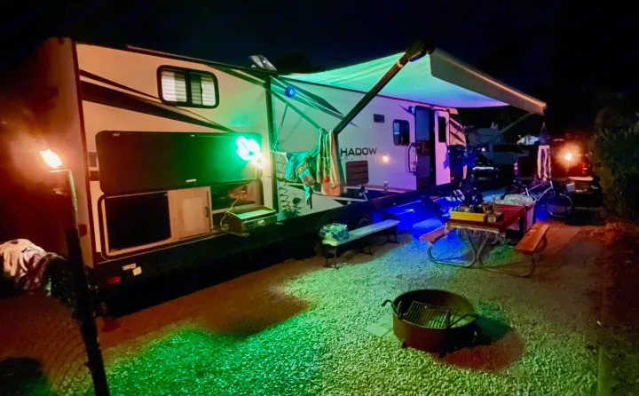 Cozy rv rental campsite at night, glowing with green & blue lights. Features outdoor kitchen, fire pit, & picnic table.. Cruiser RV Shadow Cruiser 2022