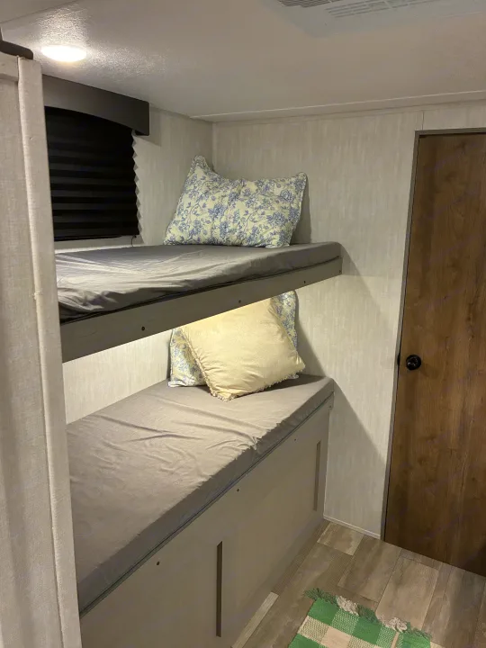 Cozy RV rental interior with bunk beds, grey linens, ceiling light, window, and wooden door.. Coachmen 25QBH 2025