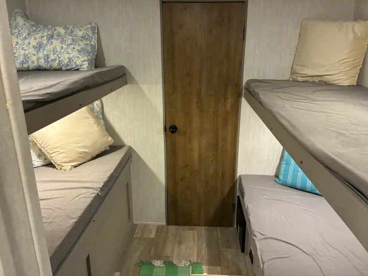 Inside an RV rental: Cozy bunk beds with grey sheets & patterned pillows, flanking a central wooden door, with a floor mat.. Coachmen 25QBH 2025