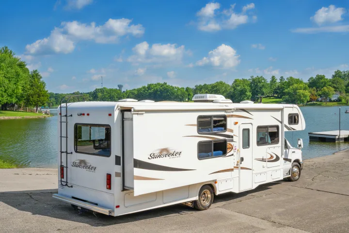 White Sunseeker RV rental parked on a boat ramp by a lake, surrounded by green trees under a blue sky.. Forest River Sunseeker 2015