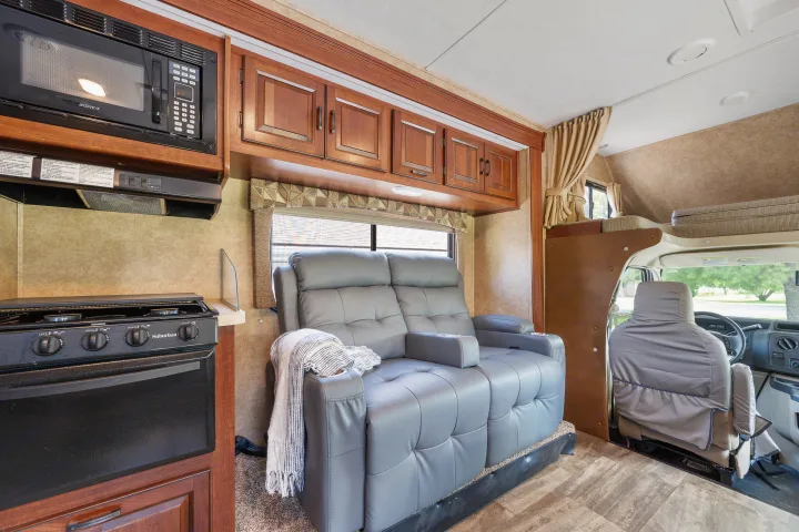 An rv rental interior showing a kitchen with microwave, Suburban stove, and a gray recliner couch. Driver's cab forward.. Forest River Sunseeker 2015