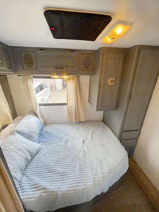 Cozy RV rental bedroom featuring a soft blue bed, window with curtains, and gray overhead cabinets.. Gulf Stream Ultra 1998