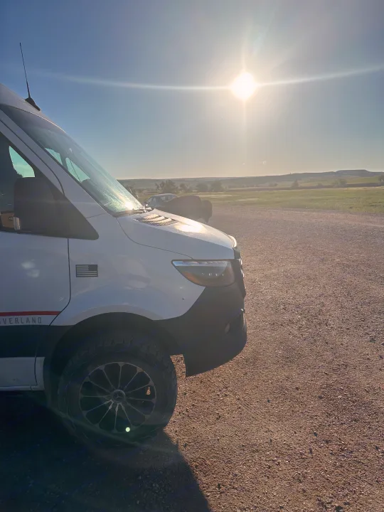 White Sprinter rv rental van on gravel road with green fields and hills under bright sun.. Storyteller Overland Storyteller Overland Class B 2021