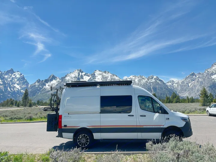 White Storyteller Overland RV rental van parked before the majestic, snow-capped Grand Teton mountains under a blue sky.. Storyteller Overland Storyteller Overland Class B 2021