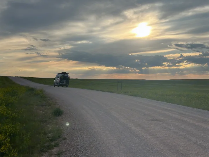Adventure-ready RV rental van on a gravel road. Sunset rays illuminate vast green fields under dramatic clouds.. Storyteller Overland Storyteller Overland Class B 2021