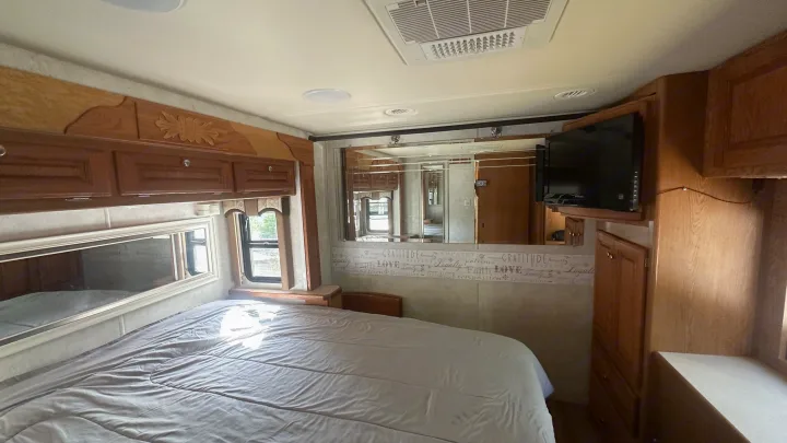 Inside this comfy RV rental bedroom: light-colored bedding on a spacious bed, wood cabinetry, TV, and inspiring wall decor.. Holiday Rambler Vacationer 2003