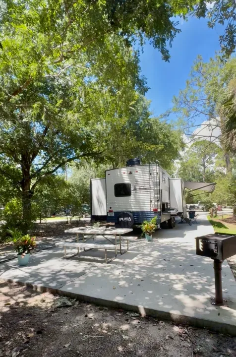 RV rental on a concrete pad at a sunny, shaded campsite with a picnic table & grill. Lush trees & blue sky.. Forest River Puma 2026