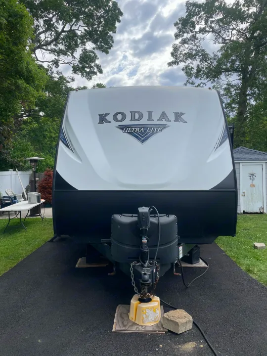 White Kodiak Ultra Lite RV rental trailer, parked on asphalt with trees and a shed visible in the background.. Dutchmen Kodiak Ultra-Light 2018