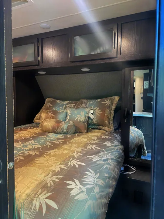 Inside an RV rental: a compact bedroom with patterned bed, overhead cabinets, and side mirror. Your travel retreat.. Dutchmen Kodiak Ultra-Light 2018