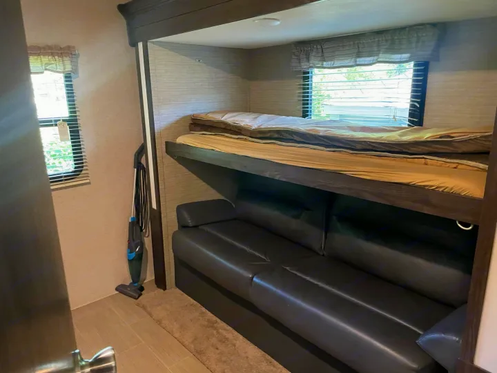 RV rental interior: upper bunk bed, lower black sofa, window, and stick vacuum.. Dutchmen Kodiak Ultra-Light 2018